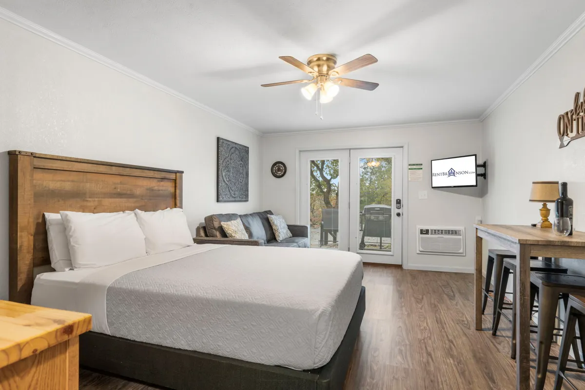 This interior shot highlights a cozy studio area with a queen bed, a soft sofa, sliding glass doors that offer plenty of natural light, a TV for streaming, and a small table with stools. The scene exudes comfort and togetherness, ideal for both relaxation and quality time.