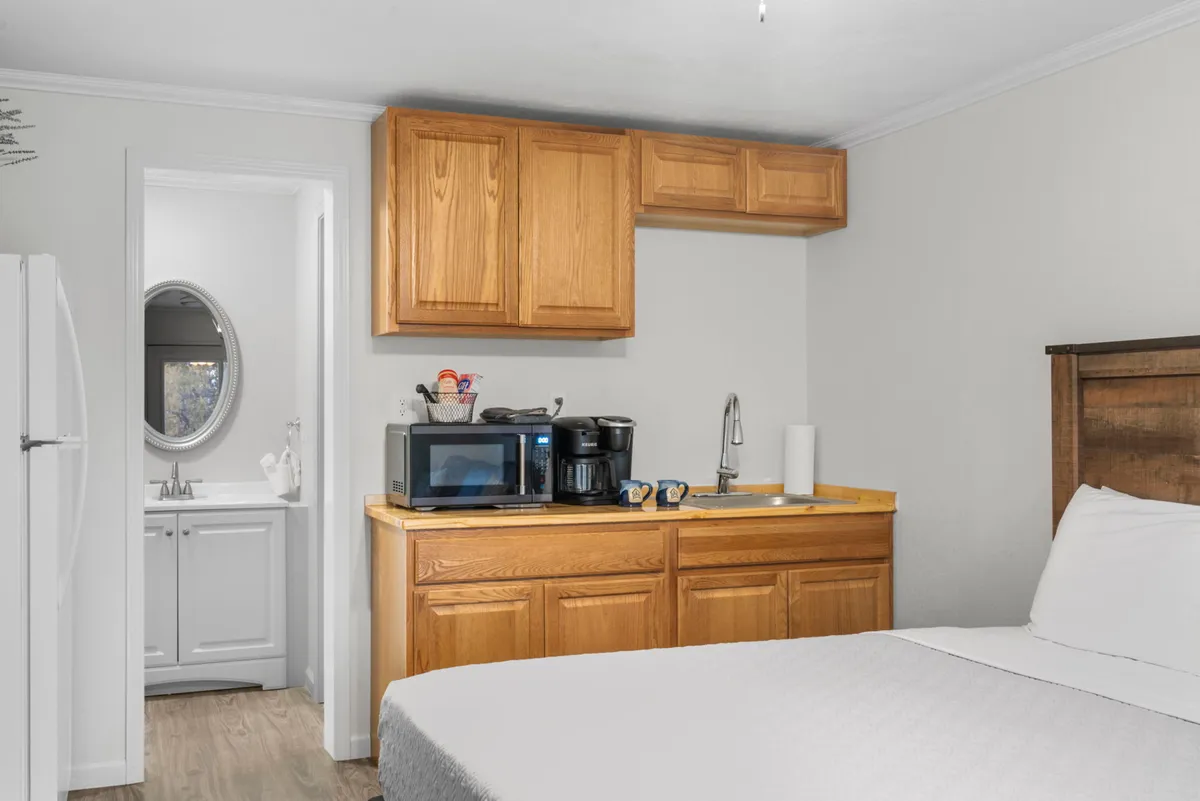 This image presents a well-lit kitchenette area integrated with a cozy sleeping space. Visible features include the compact kitchenette with cabinetry, countertop appliances, and a small refrigerator, as well as a comfortable bed and a practical mirror. The scene embodies an inviting and practical setup for everyday vacation living.