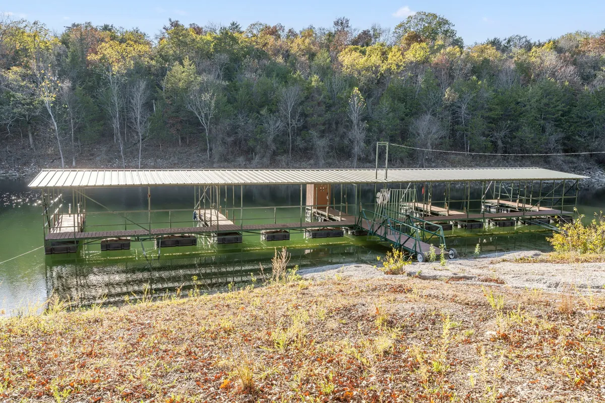 A floating dock stretches into calm, still water, set against a backdrop of leafy green trees. The setting invites activities like fishing, paddleboarding, or simply enjoying peaceful moments by the lake.