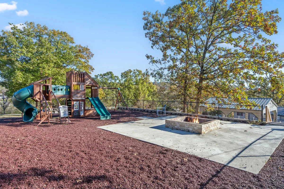 An inviting outdoor recreation space with colorful playground equipment and a fire pit under a large shady tree. Children can be seen at play while families gather and relax nearby, evoking warmth and togetherness.