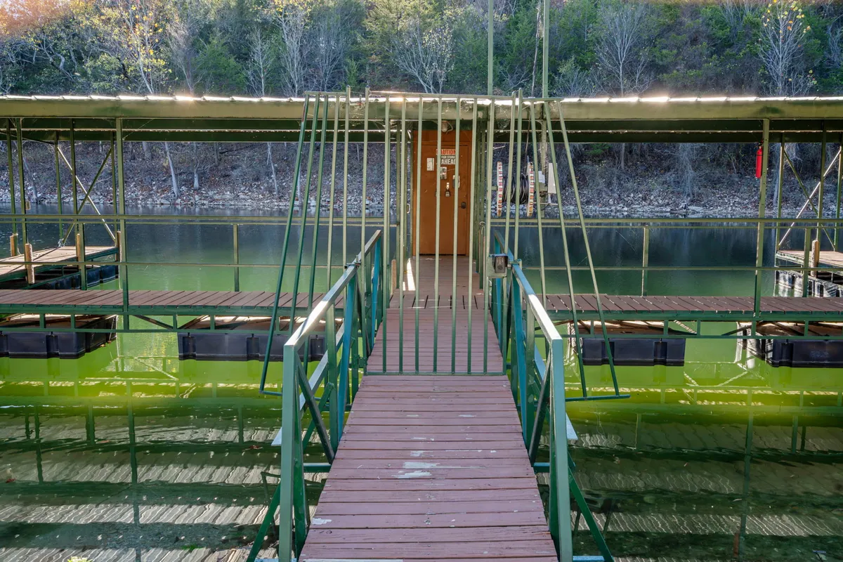 An exterior walkway with green railings leads from the condo entrance toward the water, with reflections dancing on the surface and lush trees framing the path. The peacefulness of the scene suggests a gentle transition from the home to the lakeside setting.