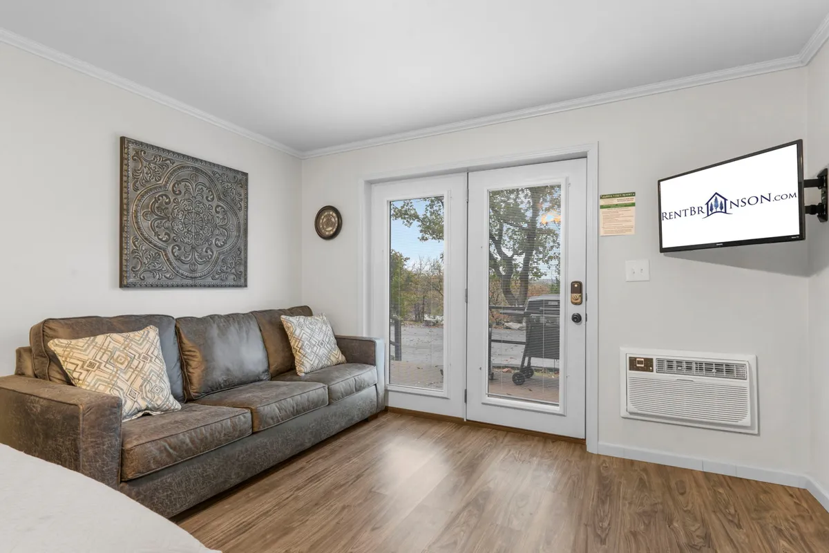 A cozy, sunlit living area with a comfortable sofa, decorative wall art, and sliding glass doors opening to a private patio. The space invites relaxation while enjoying views of the outdoors, suggesting a seamless indoor-outdoor experience.
