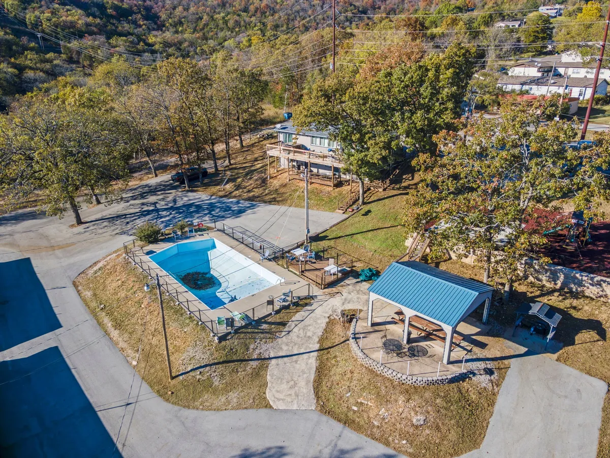 A sparkling outdoor swimming pool is framed by a gazebo, surrounded by tall trees and a nearby parking area. The scene exudes an inviting, resort-like feel, perfect for summer swimming and relaxation for all ages.