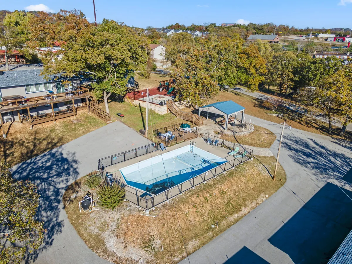 A vibrant outdoor pool area surrounded by trees, featuring a spacious deck and a shade structure. The scene captures families enjoying the pool, with space for sunbathing and gathering for barbecues. The relaxed atmosphere is perfect for both play and socializing.