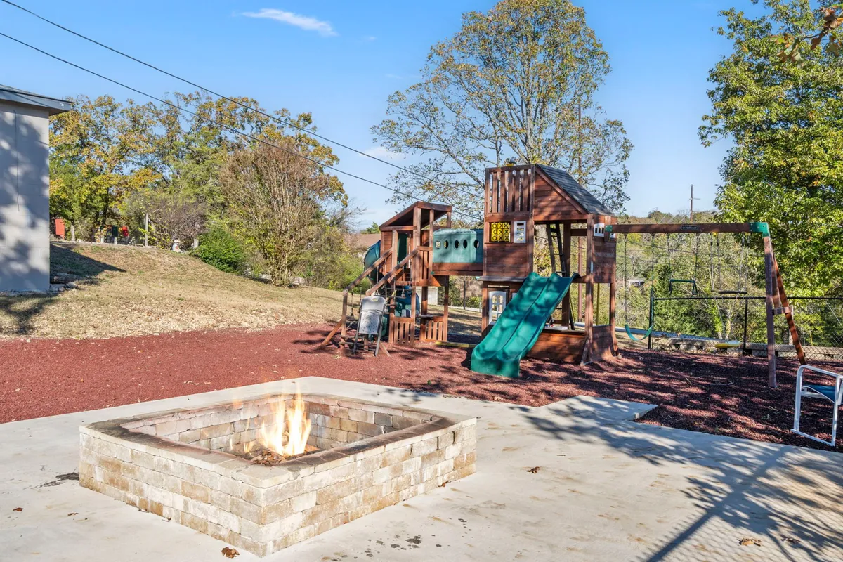 An inviting playground structure sits amid lush greenery, with a fire pit nearby and space for family gatherings. The vibrant play equipment and open area are perfect for children, while the fire pit encourages evening storytelling and marshmallow roasting.