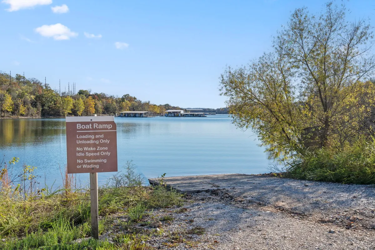 A tranquil lakeside scene shows a well-maintained boat ramp bordered by calm water and lush green trees. The area appears easily accessible, with clear signage inviting visitors to enjoy boating, fishing, or peaceful walks along the water's edge.
